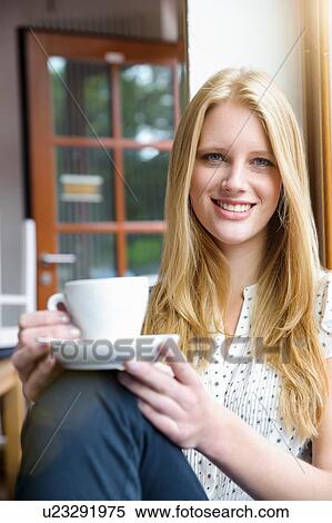 Stock Photography - "Young woman with long blond hair sitting holding coffee cup, looking at camera smiling". Fotosearch