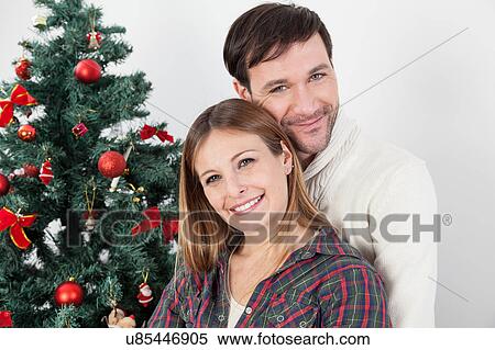 Stock Photography - Couple smiling beside the chrismas tree. Fotosearch