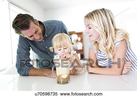 Stock Photography - Girl (2-3) drinking milkshake with parents at home. Fotosearch