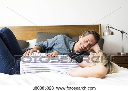 Stock Image - Husband lying down on bed with pregnant wife. Fotosearch