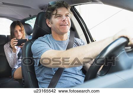 Stock Image - Man driving car, girl (10-11) texting in background. Fotosearch