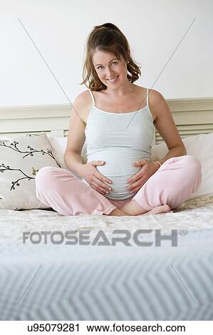 Stock Image - Smiling pregnant woman sitting on bed. Fotosearch