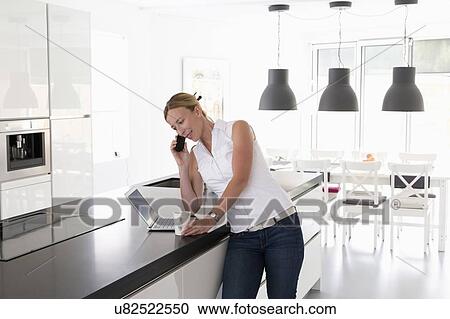 Stock Image - Woman with laptop in kitchen talking on phone. Fotosearch