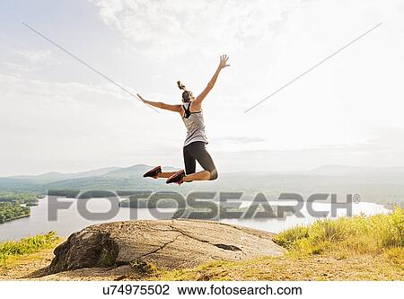 Stock Image - Young woman jumping over mountain top, rear view. Fotosearch