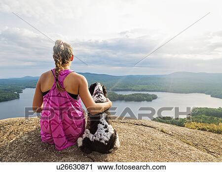 Stock Image - Young woman sitting with dog on cliff looking at lake view. Fotosearch