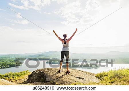 Stock Image - Young woman standing on top of mountain with outstretched arms, rear view. Fotosearch