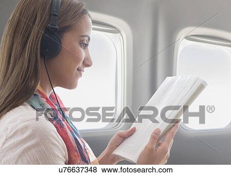 Young woman wearing headphones while reading book on plane View Large Photo Image Stock Photo - Young woman wearing headphones while reading book on plane. Fotosearch