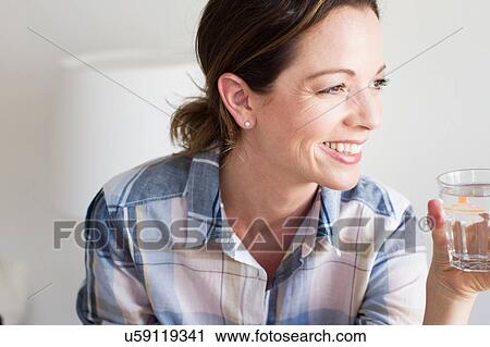 Stock Image - Cropped view of mature woman holding glass of water looking away smiling. Fotosearch