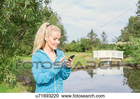 Stock Image - Female runner selecting earphone music on smartphone in park. Fotosearch