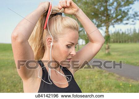 Stock Photograph - Female runner with smartphone tying pony tail in park. Fotosearch