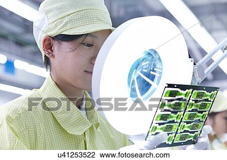 Stock Image - Female worker using magnifying glass at quality check station for a factory producing flexible electronic circuit boards. Plant is located in the south of China, in Zhuhai, Guangdong province. Fotosearch