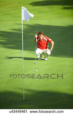Stock Image - Front view of golfer crouching down in front of golf flag considering stategy. Fotosearch