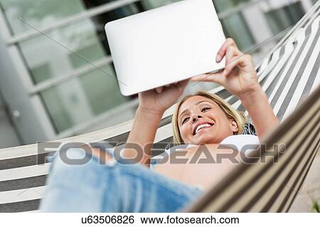 Stock Photograph - Full term pregnancy young woman lying in hammock using digital tablet. Fotosearch