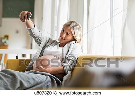 Stock Photo - Full term pregnancy young woman on sofa taking selfie. Fotosearch