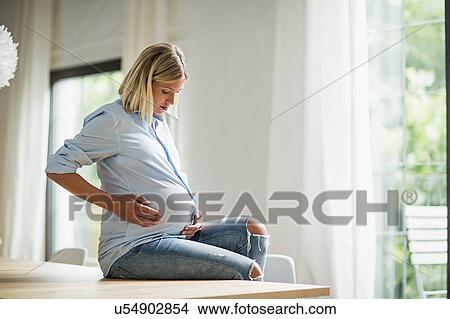 Picture - Full term pregnancy young woman sitting on kitchen table. Fotosearch