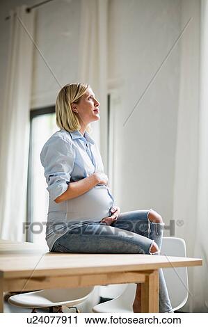 Stock Image - Full term pregnancy young woman sitting on table holding stomach. Fotosearch
