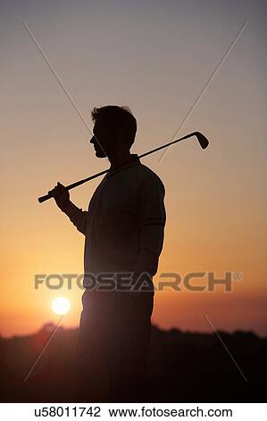 Stock Image - Golfer carrying golf club over shoulder in front of sunset, looking away. Fotosearch