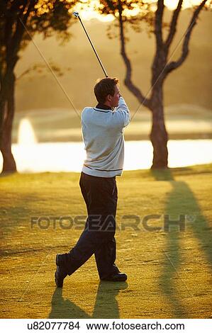 Picture - Golfer in sunlight holding golf club taking golf swing, looking away. Fotosearch