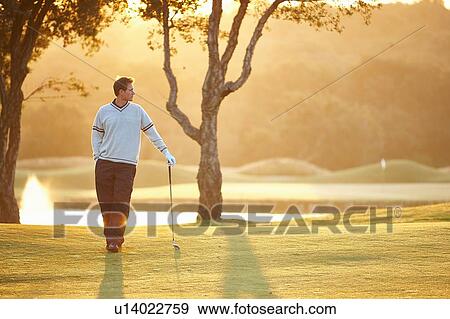 Stock Photo - Golfer in sunlight leaning against golf club, hand in pocket looking away. Fotosearch
