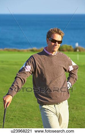 Stock Image - Golfer wearing sunglasses leaning against golf club, hand on hip, looking at camera. Fotosearch
