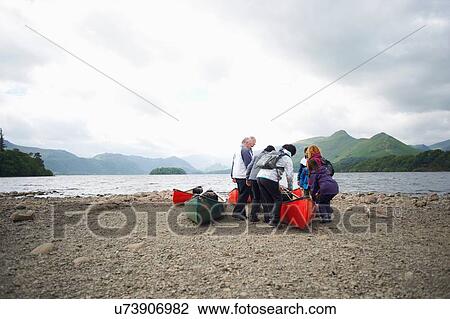 Stock Image - Group of people standing by canoe at lake edge, Lake District, Cumbria, UK. Fotosearch