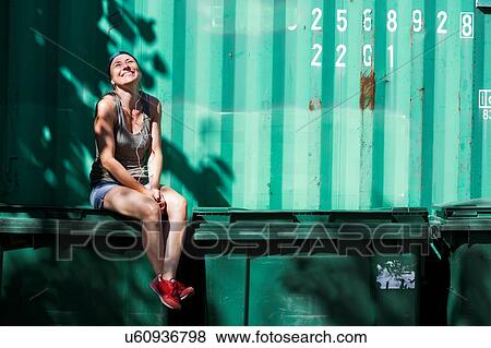 Stock Photo - Happy woman sitting on recycle bin beside green container. Fotosearch