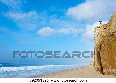 Stock Photography - Low angle view of golder on cliff top overlooking beach taking golf swing. Fotosearch