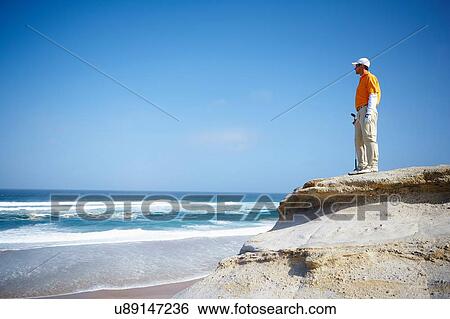 Stock Photograph - Low angle view of golfer standing on cliff overlooking ocean looking away. Fotosearch