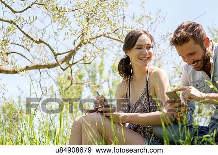 Low angle view of young couple sitting looking down at smartphone smiling View Large Photo Image Stock Photo - Low angle view of young couple sitting looking down at smartphone smiling. Fotosearch