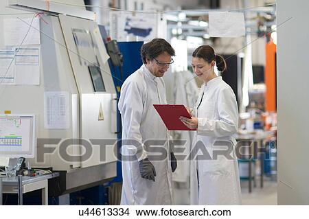 Picture - Male and female colleagues wearing labcoats looking at clipboard smiling. Fotosearch