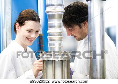 Stock Image - Male and female colleagues wearing labcoats using scientific equipment, looking down smiling. Fotosearch