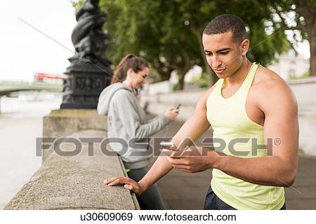 Stock Photo - Male and female runners reading smartphone texts on riverside. Fotosearch