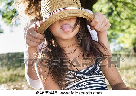 Stock Photo - Mature woman covering daughters eyes with straw hat in park. Fotosearch