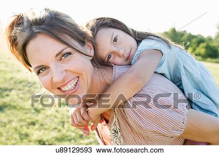 Stock Image - Mature woman giving daughter piggy back ride in park. Fotosearch