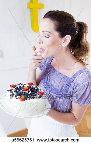 Stock Image - Mature woman holding fruit covered cake, licking fingers eyes closed. Fotosearch