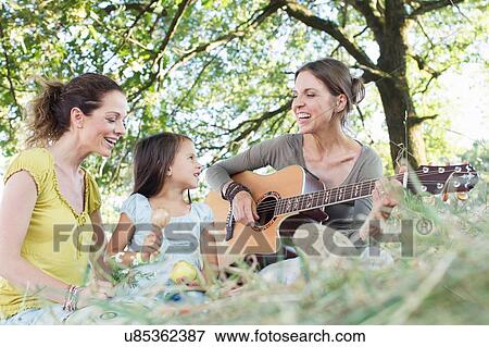 Stock Photo - Mature woman playing acoustic guitar for friend and daughter in park. Fotosearch