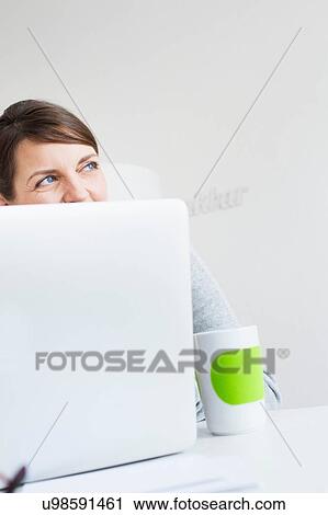 Stock Image - Mature woman sitting behind laptop, face obscured, looking away. Fotosearch