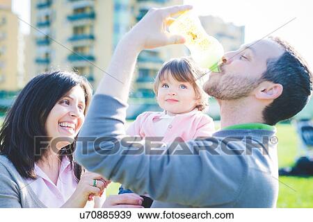 Stock Photo - Mid adult man drinking toddler daughters baby bottle in park. Fotosearch