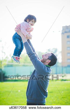 Stock Photo - Mid adult man lifting up toddler daughter in park. Fotosearch