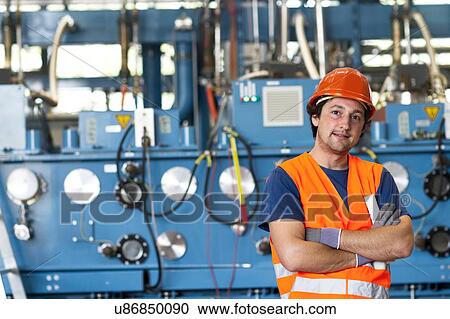 Stock Image - Mid adult man wearing hard hat and safety vest in front of scientific equipment, arms folded looking at camera. Fotosearch