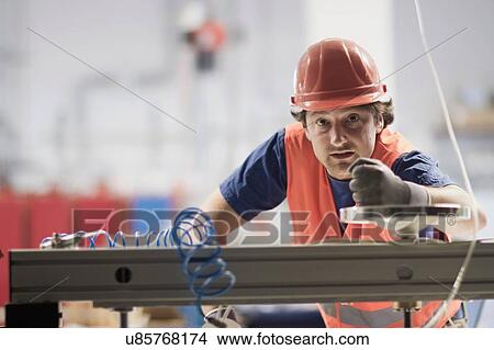 Picture - Mid adult man wearing hard hat looking at camera, operating machine. Fotosearch