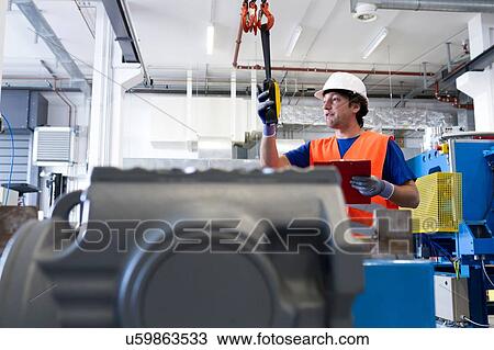 Stock Image - Mid adult man wearing hard hat operating machine, holding clipboard. Fotosearch
