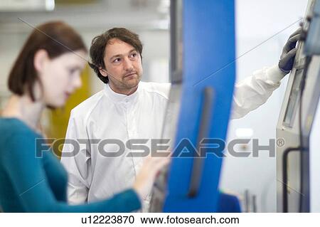 Stock Image - Mid adult man wearing labcoat and protective gloves operating machine, looking away. Fotosearch