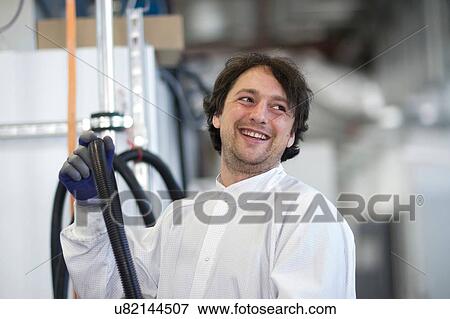 Stock Photo - Mid adult man wearing labcoat holding industrial piping, looking away smiling. Fotosearch