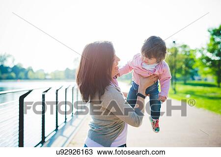 Stock Image - Mid adult woman lifting up toddler daughter at riverside. Fotosearch