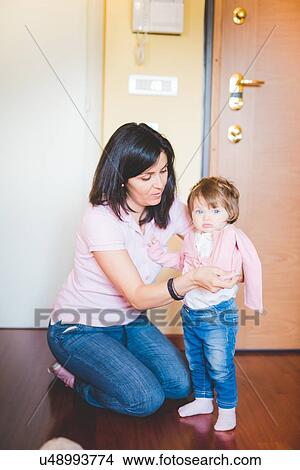 Picture - Mid adult woman putting on toddler daughters cardigan. Fotosearch