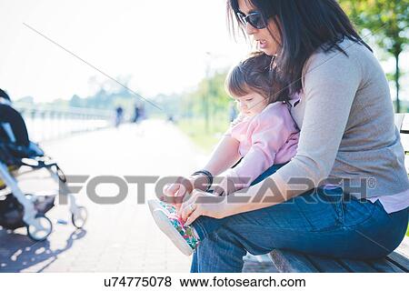 Stock Photo - Mid adult woman tying toddler daughter shoelaces in park. Fotosearch