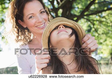 Stock Photo - Portrait of mature woman covering daughters eyes with straw hat in park. Fotosearch