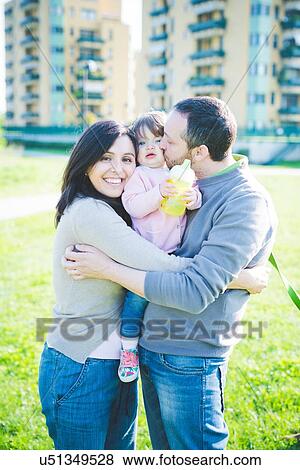 Stock Photo - Portrait of mid adult couple kissing toddler daughter in park. Fotosearch