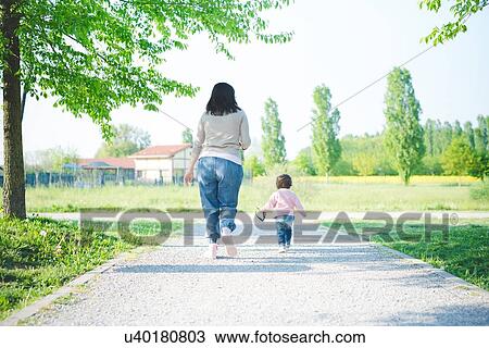 Stock Image - Rear view of female toddler toddling with mother in park. Fotosearch